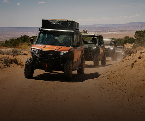A convoy of Polaris XPEDITION Side-By-Side vehicles navigates a sandy desert trail, with the lead orange vehicle featuring a roof tent and bright LED light bar.
