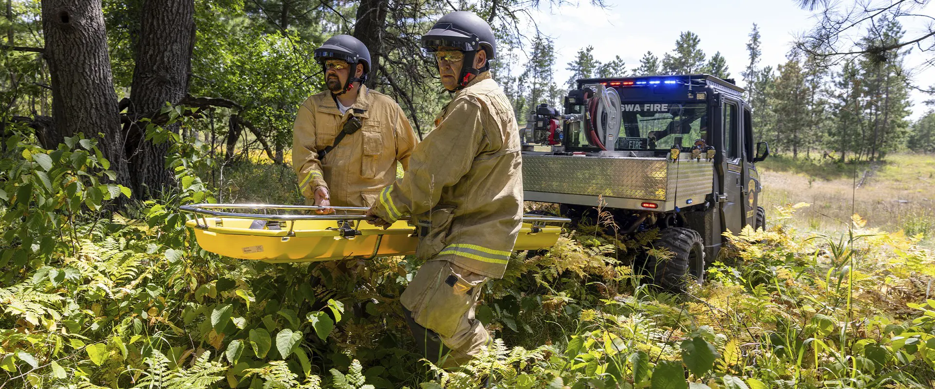 Two first responders carry a rescue stretcher from a Polaris RANGER utility vehicle in dense Minnesota woods.