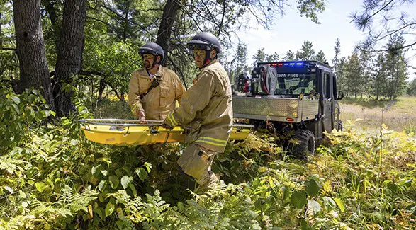 Two first responders carry a rescue stretcher from a Polaris RANGER utility vehicle in dense Minnesota woods.