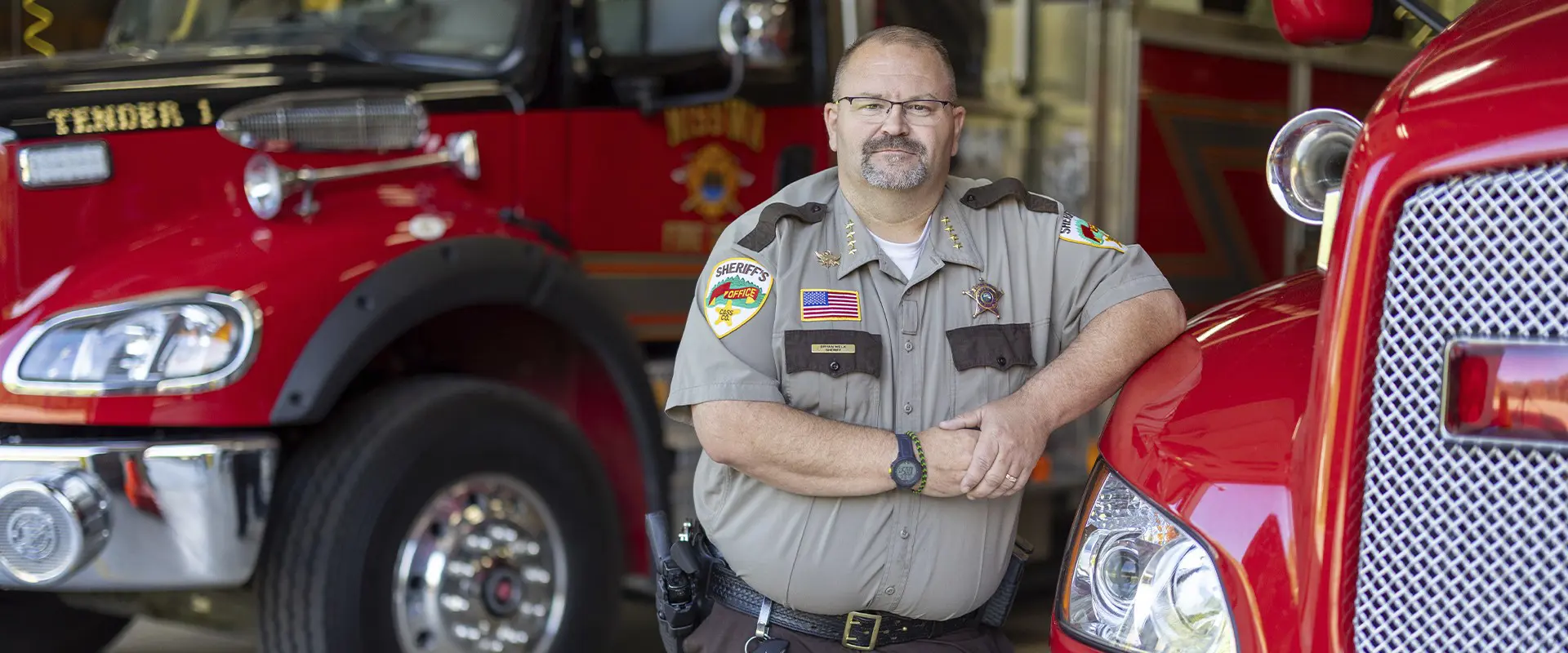 Cass County sheriff’s officer standing between two red fire trucks inside a fire station.