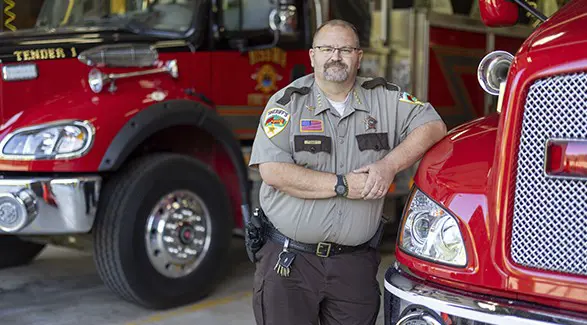 Cass County sheriff’s officer standing between two red fire trucks inside a fire station.