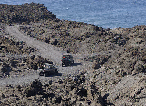 Two Polaris RANGER UTVs driving along a winding dirt trail through rocky coastal terrain, with rugged volcanic rocks in the foreground and ocean visible in the background.