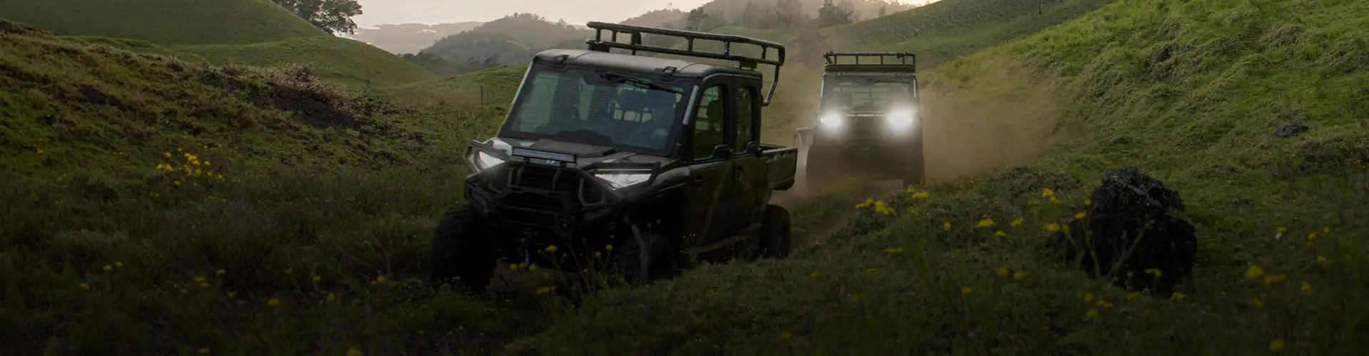Two Polaris RANGER UTVs, featuring a lead CREW model, navigate a grassy Maui hillside with headlights on at dusk, kicking up dust.