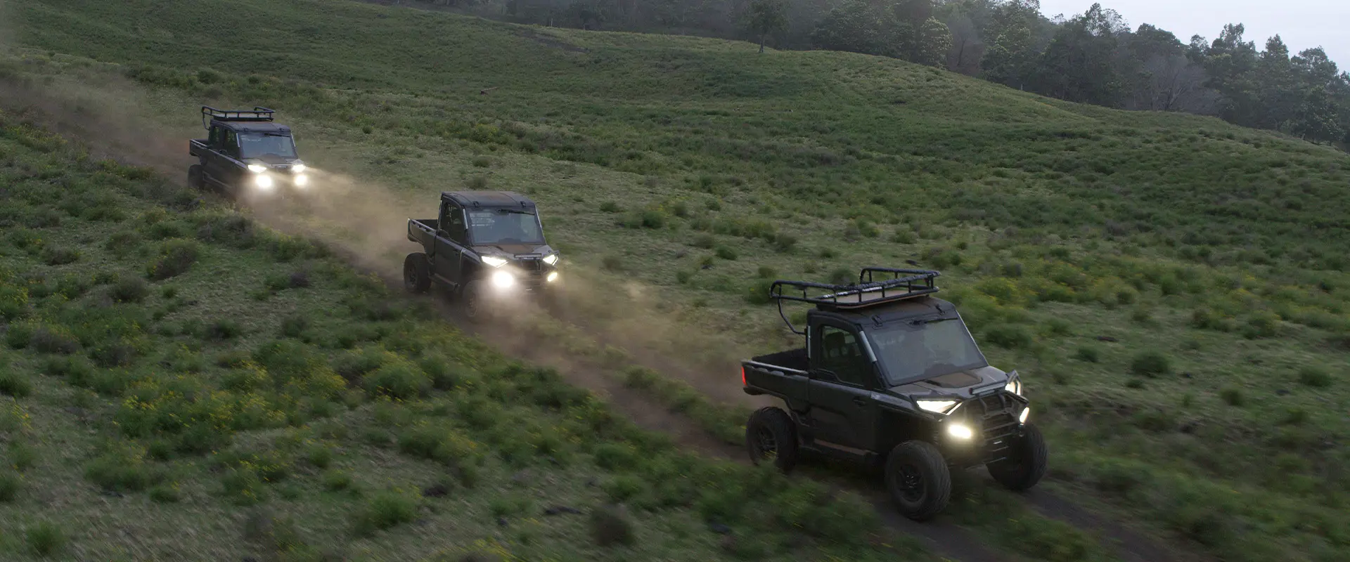 A convoy of black Polaris RANGER UTVs, including a 6-Seat model, navigates a grassy Maui hillside with headlights on at dusk.