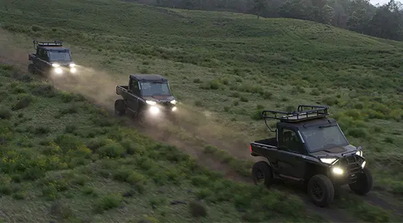 A convoy of black Polaris RANGER UTVs, including a 6-Seat model, navigates a grassy Maui hillside with headlights on at dusk.
