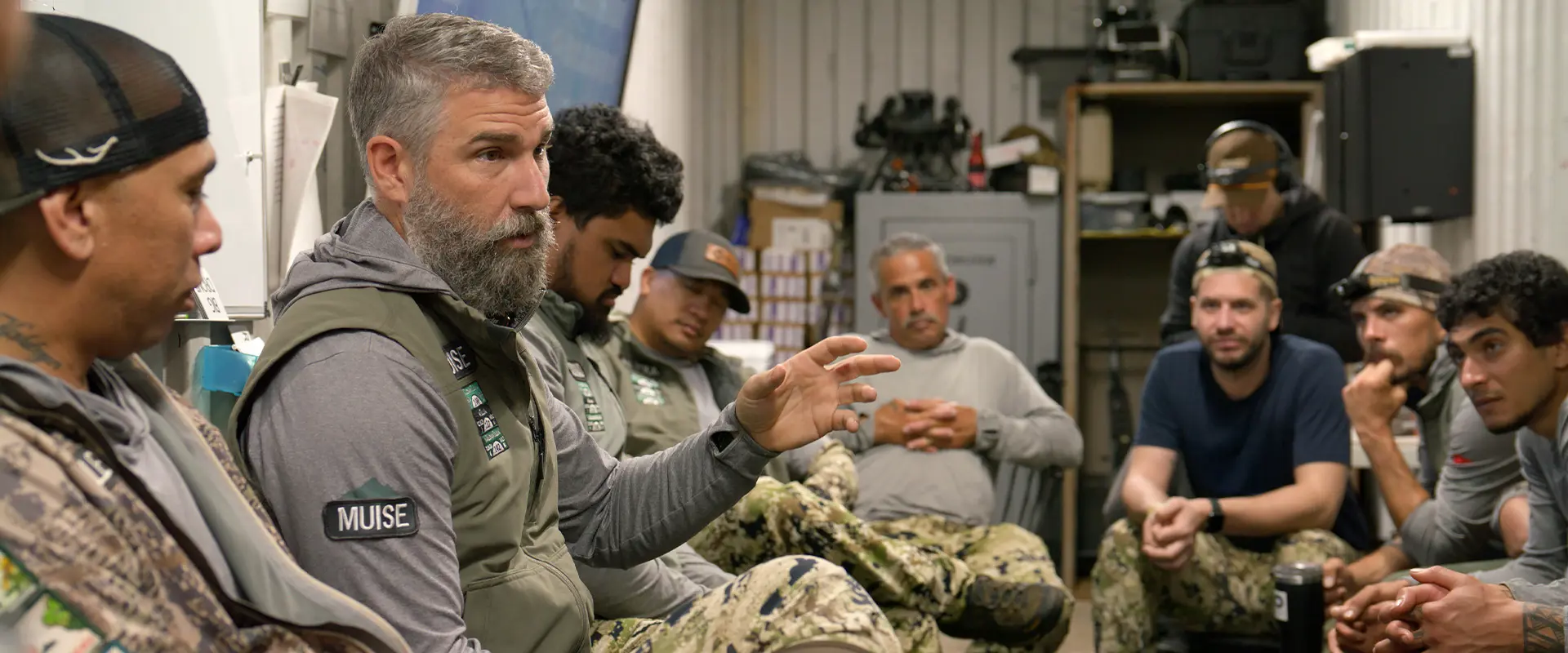 A team of men in camouflage gear listens to a mission debrief for Maui Nui Venison led by a man with a grey beard.