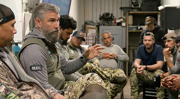 A team of men in camouflage gear listens to a mission debrief for Maui Nui Venison led by a man with a grey beard.