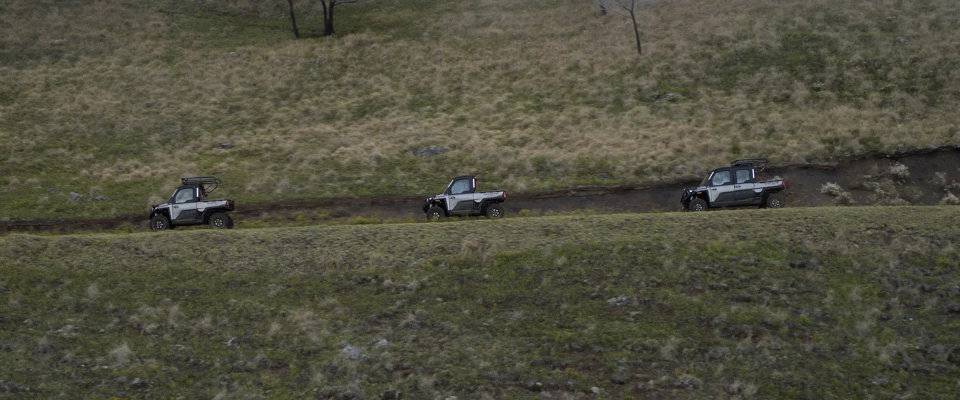 Three Polaris RANGER UTVs, including a CREW model, traverse a high-elevation dirt road along a vast, grassy ridge.