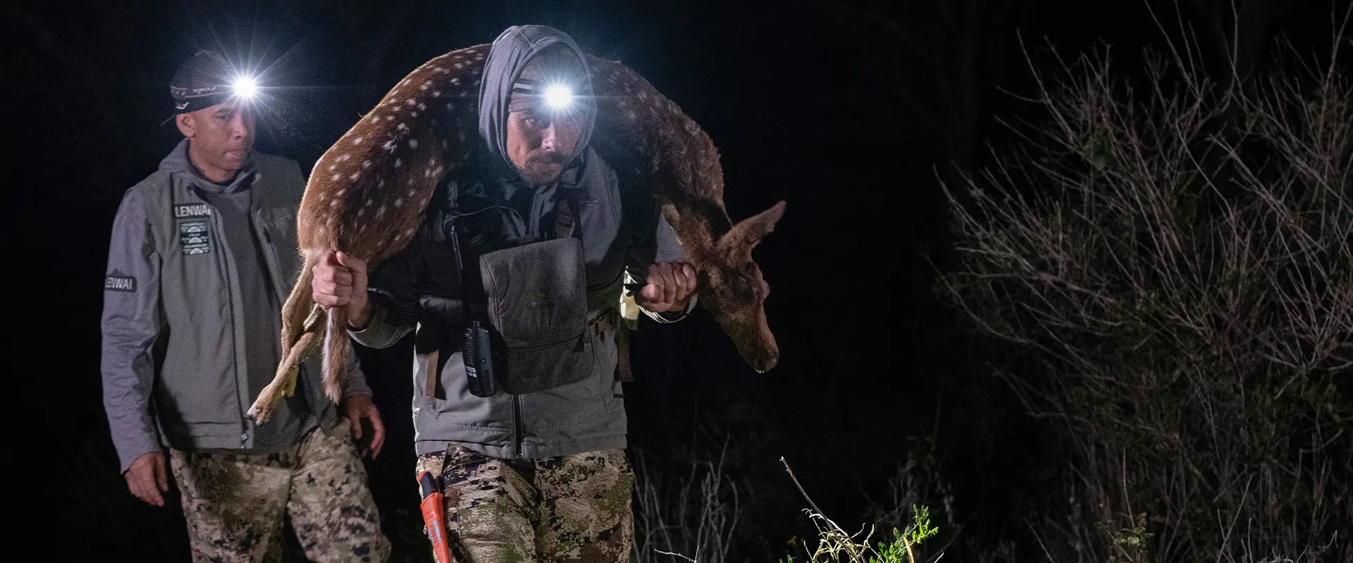 Two men using headlamps harvest venison at night, with one carrying a deer over his shoulders through the dark brush.