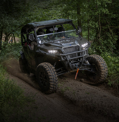 Polaris Off Road Vehicle drives through a dense forest trail, navigating muddy terrain with headlights on and tall greenery surrounding the narrow path.