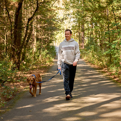A man walking a dog along a wooded trail while wearing a gray Polaris hoodie and black joggers.
