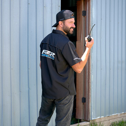 A man standing beside a building entrance wearing a Polaris RZR branded work shirt and cap while holding a two-way radio.