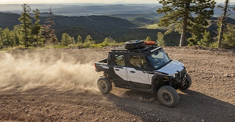 Polaris XPEDITION XP 5 NorthStar SxS driving on a rocky mountain trail, kicking up dust while carrying overlanding gear on the roof rack above a forested landscape.