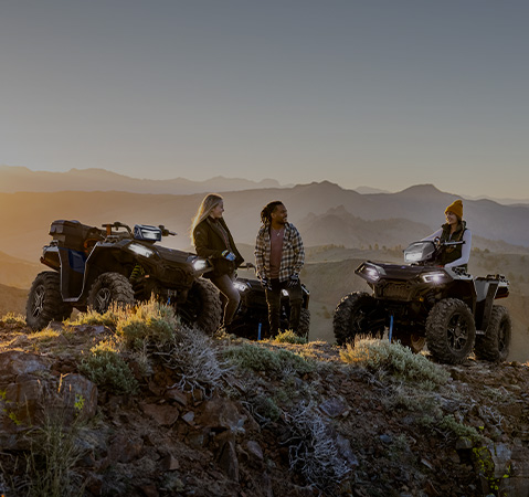 Tres amigos de pie junto a dos cuatrimotos Polaris Sportsman en una cresta montañosa con vistas panorámicas al atardecer.