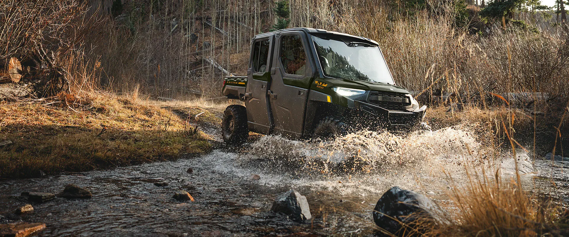 A Polaris RANGER CREW XP 1000 NorthStar enclosed cab UTV driving through a shallow river on a summer day.