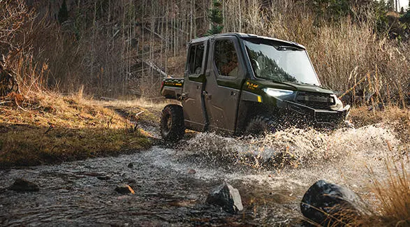 A Polaris RANGER CREW XP 1000 NorthStar enclosed cab UTV driving through a shallow river on a summer day.