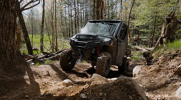 A Polaris RANGER XP 1000 NorthStar enclosed cab UTV climbing a rocky hill in the forest on a summer day.