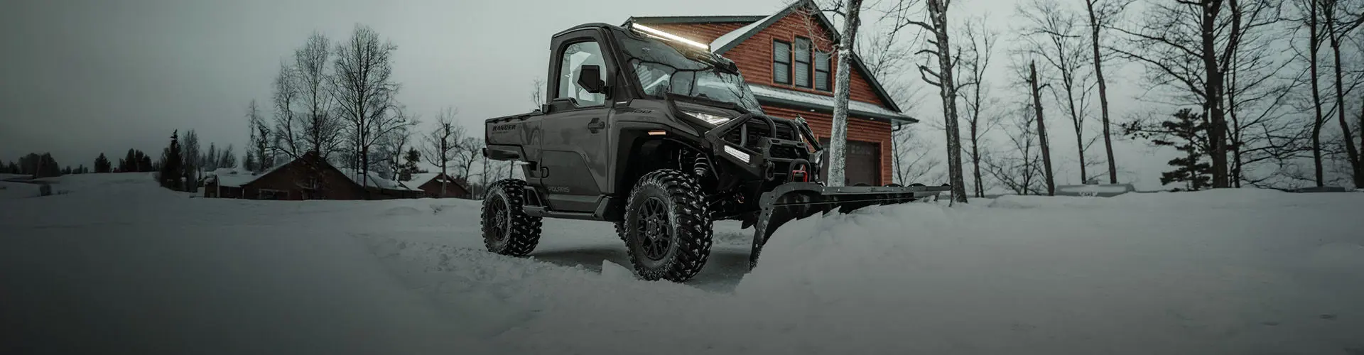 A Polaris RANGER UTV with a snow plow clears snow in front of a wooden house on a snowy day.