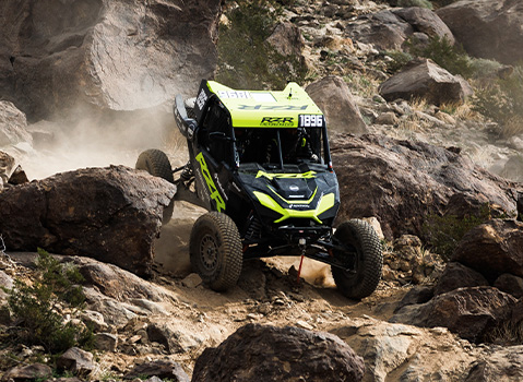 A black and lime green Polaris RZR Factory Racing Side-by-Side vehicle climbs over massive boulders on a rugged, dusty trail.