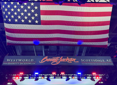 A large American flag hangs above a Barrett-Jackson Scottsdale auction banner at WestWorld in Arizona, with red and blue stage lights below.