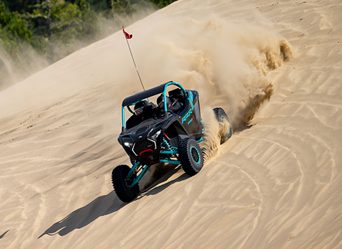 A black and turquoise Polaris RZR Pro R Side-by-Side kicks up a massive cloud of sand while climbing a steep dune.