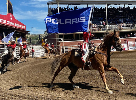 Riders on horseback carry a large blue Polaris flag during a performance in a crowded outdoor arena.