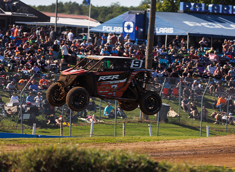 A red and black Polaris RZR SxS race vehicle catches air as it jumps over a dirt track during an off-road competition.