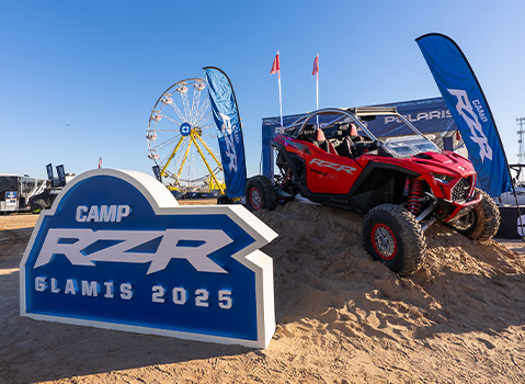 A red Polaris RZR Pro R SxS is displayed on a sand mound next to the Camp RZR Glamis 2025 sign with a Ferris wheel behind it.