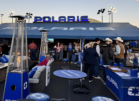 People gather at a Polaris outdoor event booth featuring blue branded furniture and pyramid heaters at dusk.