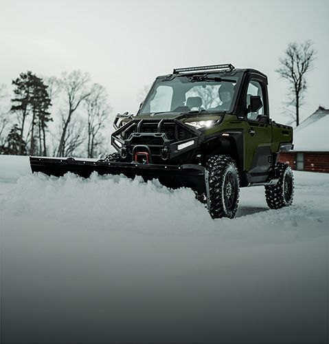 A dark green Polaris RANGER UTV with a snow plow attachment clears snow in a winter landscape with trees and a cabin in the background.