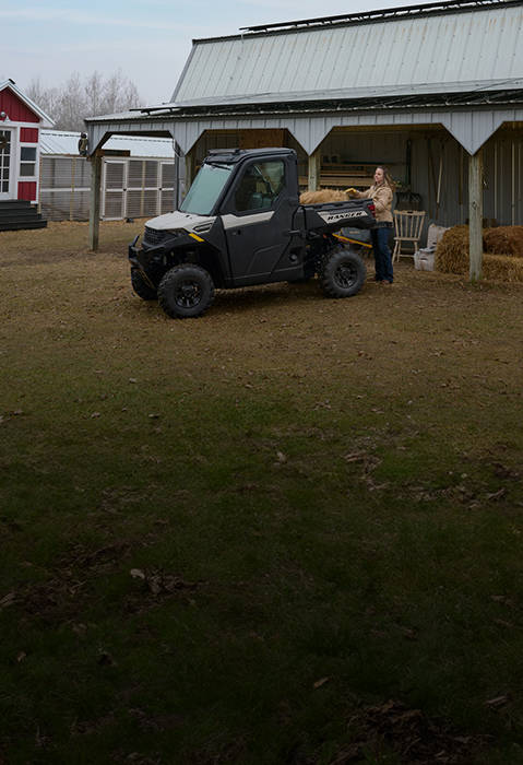 A Polaris Ranger 1000 UTV with two people in helmets and sunglasses drives on a dirt road, carrying tools in its cargo bed, with fields and trees in the background.