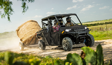 A 2026 Polaris RANGER 1000 UTV, with four passengers wearing helmets, kicks up dust as it pulls a trailer loaded with a large hay bale.