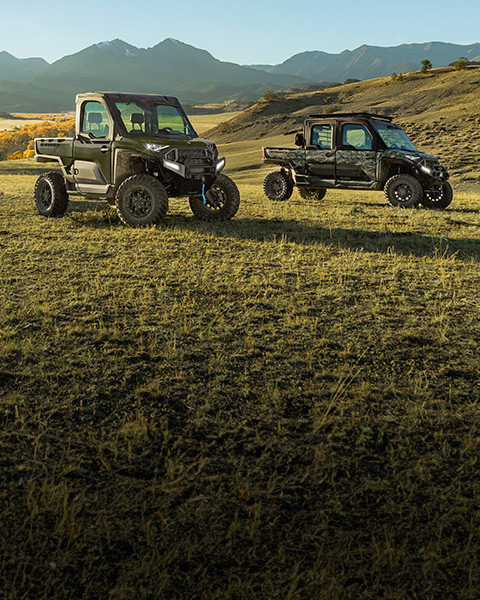 Dos UTV Polaris RANGER XD 1500 NorthStar Edition, uno verde oliva y otro en colores de camuflaje, están estacionados en una ladera con césped y montañas ondulantes de otoño en el fondo.