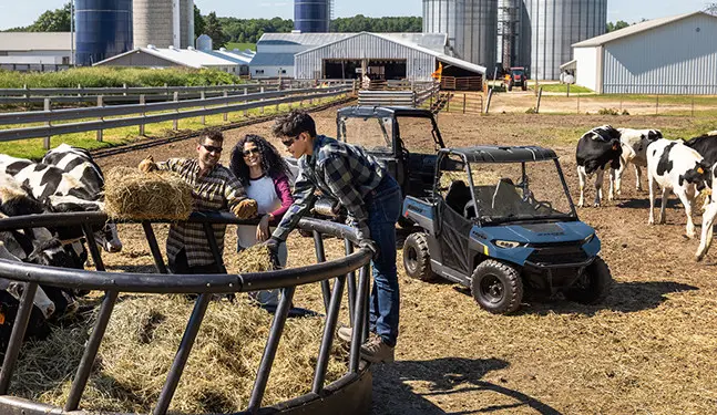 A kid standing on a bale ring with his 2027 Polaris RANGER 150 EFI youth UTV in the background near cows.