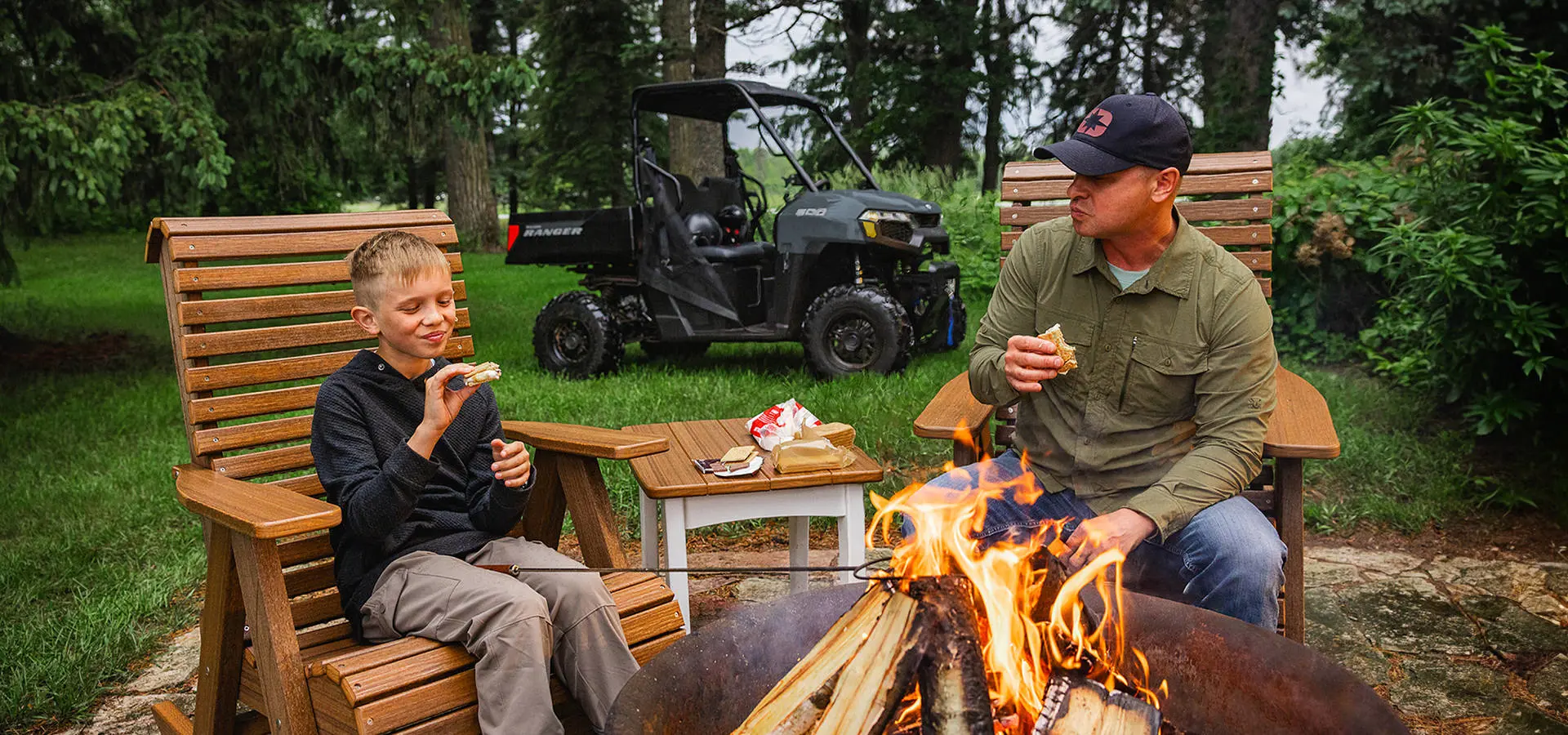 A man and his son having smores by a fire while their 2027 Polaris RANGER 500 UTV is parked behind them.