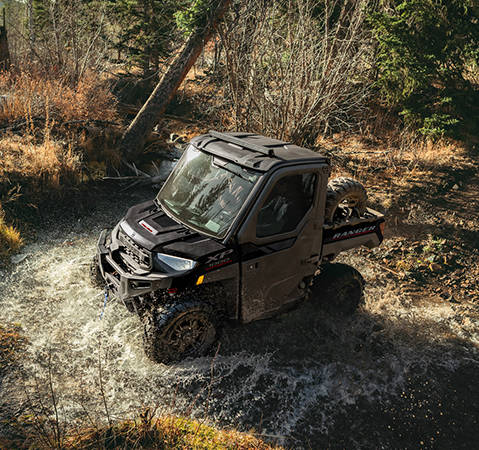 2 riders in the factory-installed cab of a 2027 Polaris RANGER XP 1000 NorthStar Trail Boss Edition UTV while driving through a ravine.
