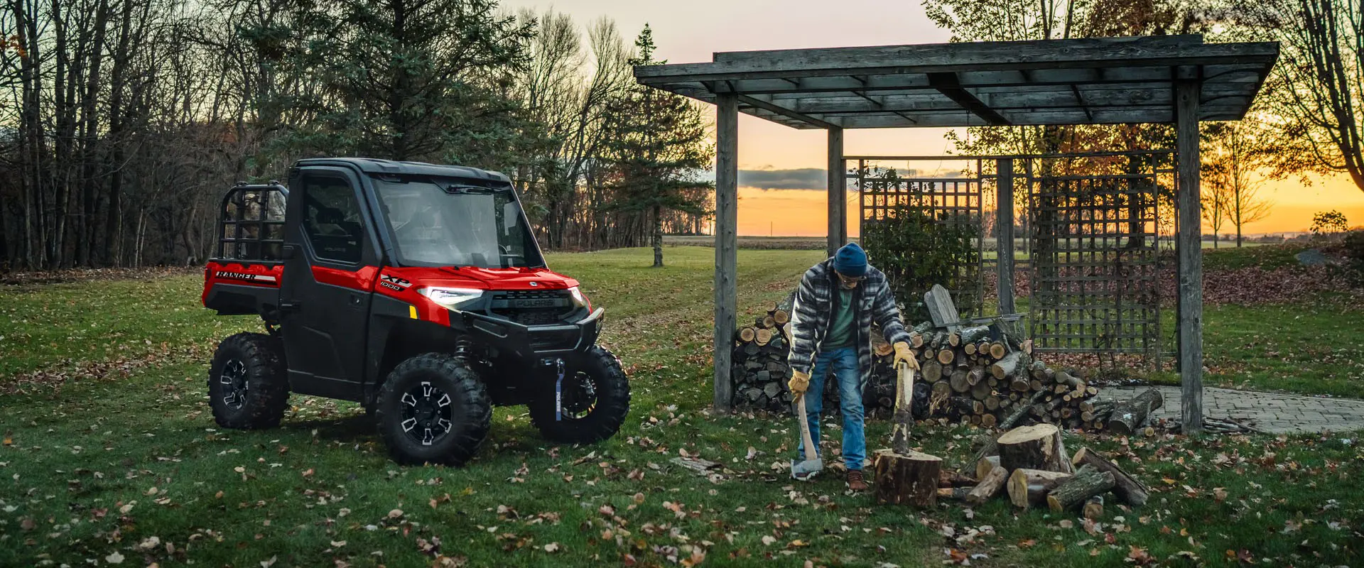 A man cutting wood at dusk illuminated by the headlights from his 2027 Polaris RANGER XP 1000 NorthStar enclosed UTV.