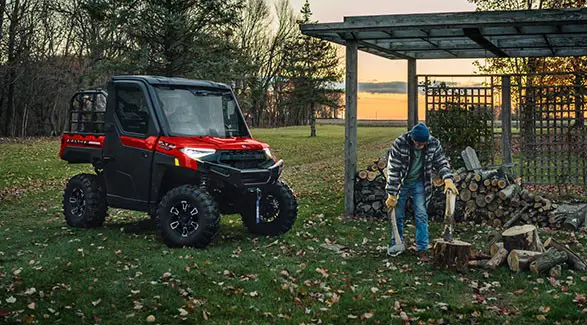 A man cutting wood at dusk illuminated by the headlights from his 2027 Polaris RANGER XP 1000 NorthStar enclosed UTV.