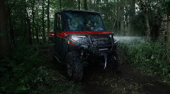 2 riders in a 2027 Polaris RANGER XP 1000 NorthStar enclosed UTV driving through a forest in the rain at night.