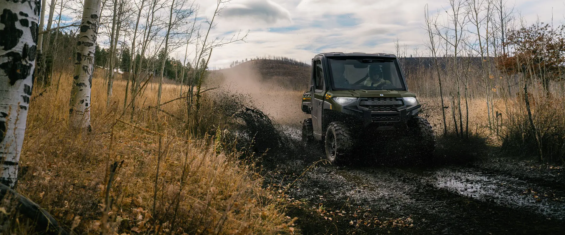 2 riders in a 2027 Polaris RANGER XP 1000 NorthStar enclosed UTV driving through a muddy section of trail.