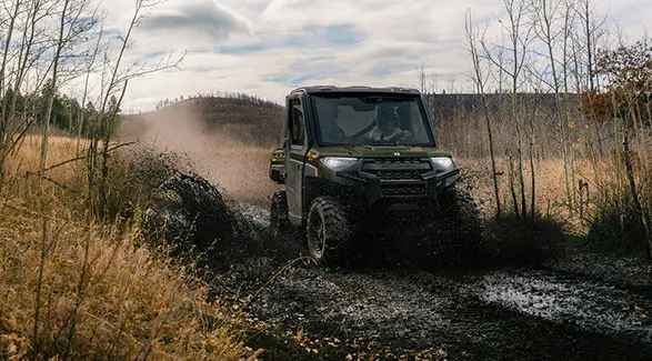 2 riders in a 2027 Polaris RANGER XP 1000 NorthStar enclosed UTV driving through a muddy section of trail.