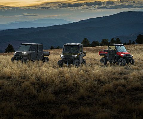 Three Polaris RANGER utility vehicles parked in a grassy mountain landscape at dusk.