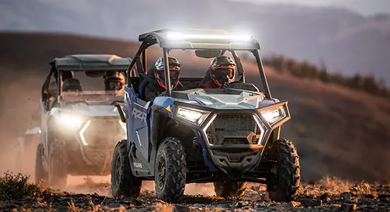 un RZR trail sport qui traverse un sentier rocheux, avec ses phares allumés. suivi d’un autre RZR