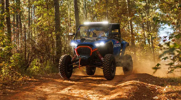 A blue 2025 Polaris RZR XP with illuminated headlights on a forest trail.