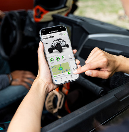 A person is holding a smartphone displaying the Polaris RIDE COMMAND app, showing vehicle information, ride details, and a map with a Location Alarm toggle.