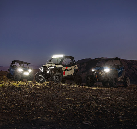 Three 2025 Polaris RZR Trail S and RZR Trail side-by-sides with their headlights and lightbars illuminated at night. 