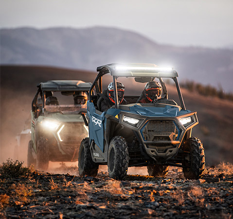 Two 2025 RZR Trail side-by-sides cruising with LED lighting accessories installed.