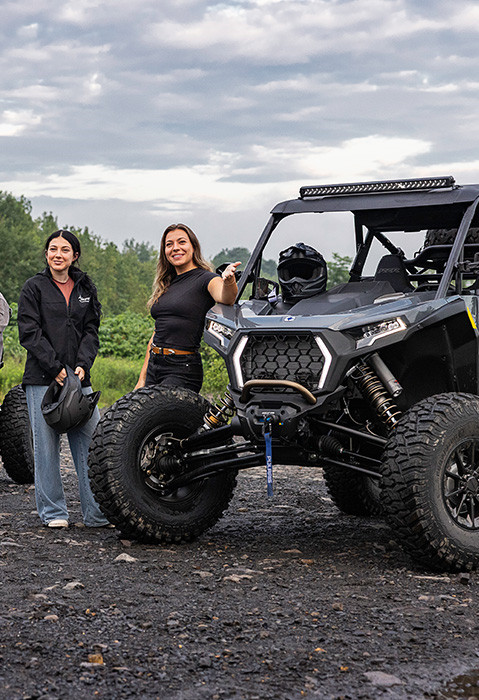 Polaris RZRs with roll cages, light bars, and custom suspension systems in a desert terrain with rocky hills, featuring riders interacting with the SxS.
