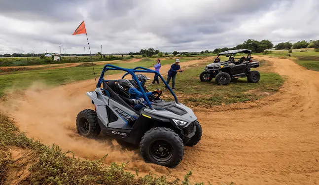 Side view of 2 kids in a 2026 Polaris RZR 200 EFI youth SxS driving on an open course.