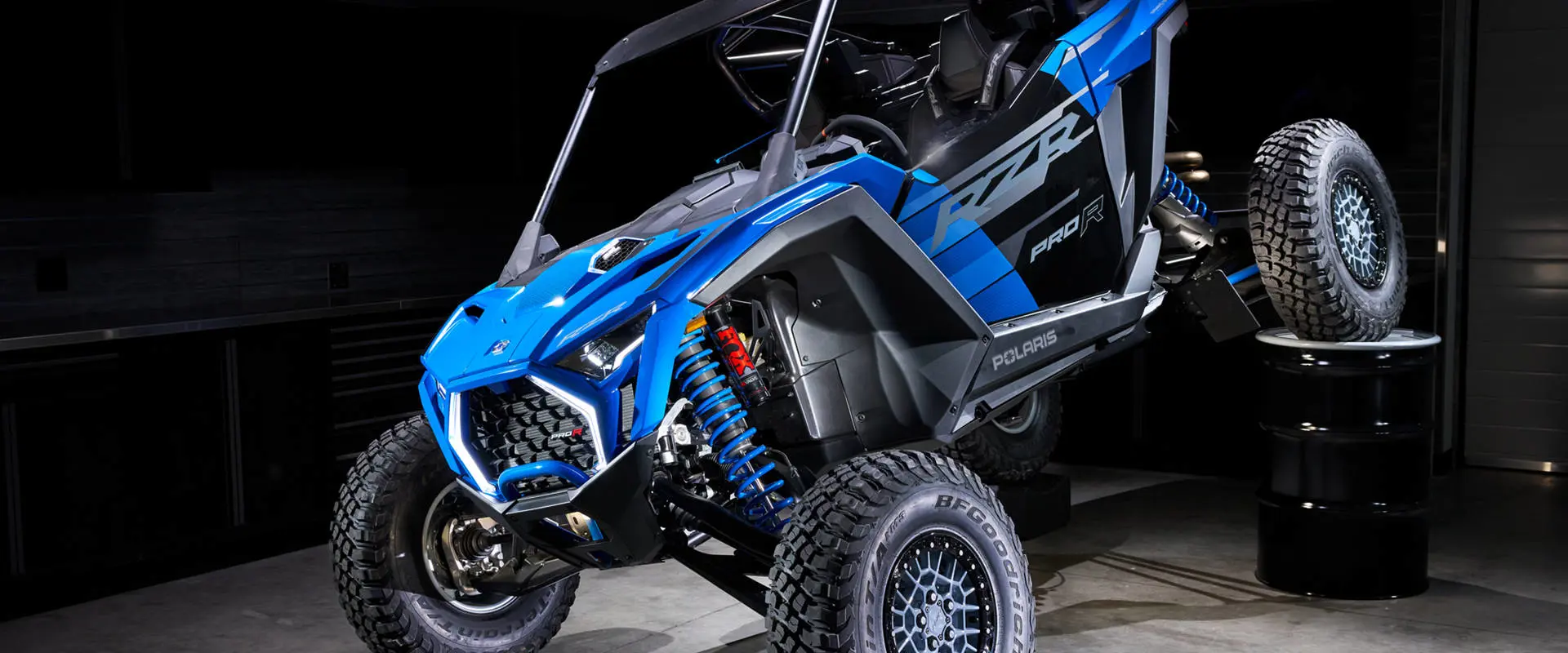 A front-angled shot of a Polaris RZR Pro R Ultra Edition Velocity Blue with one tire propped on a barrel to highlight its FOX suspension.